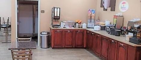 A small kitchenette with a coffee machine, a trash can, and wooden cabinets.
