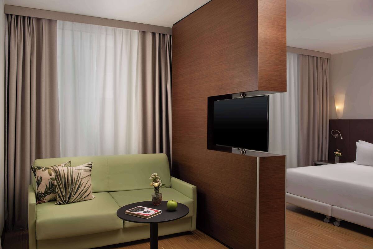 A hotel room with a green sofa, a small table with a book and a vase, a bed, and a flat-screen TV mounted on the wall.
