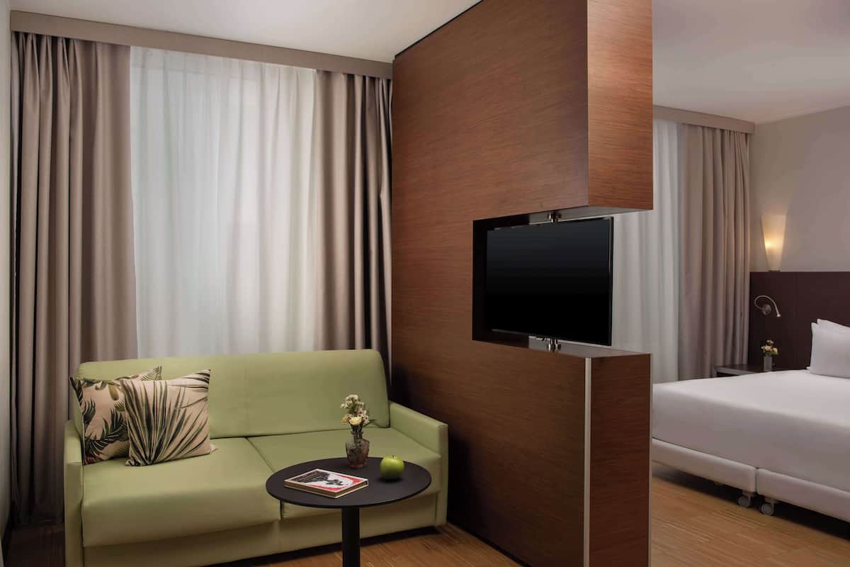 A hotel room with a green sofa, a small table with a book and a vase, a bed, and a flat-screen TV mounted on the wall.