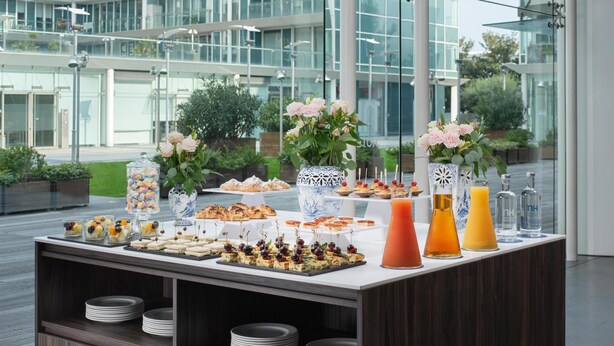 A buffet table with various food items and drinks, set against a modern building with large windows.