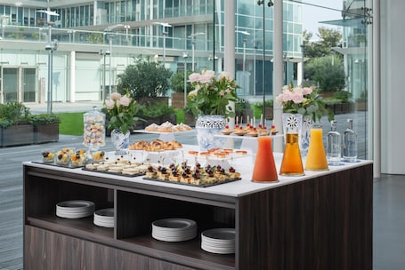 A buffet table with various food items and drinks, set against a modern building with large windows.