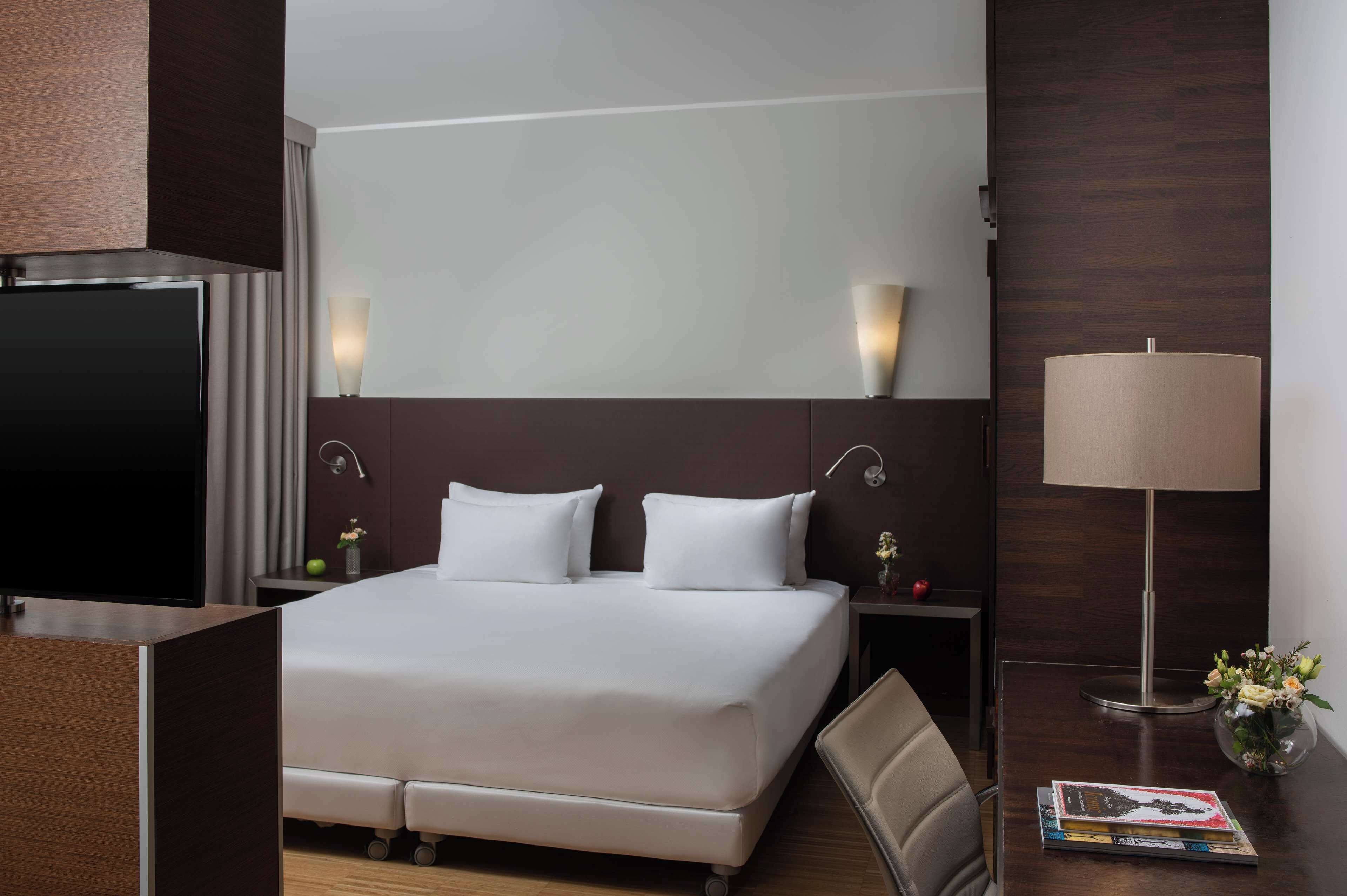 A hotel room with a large bed, a TV, a desk, a lamp, and a vase with flowers.