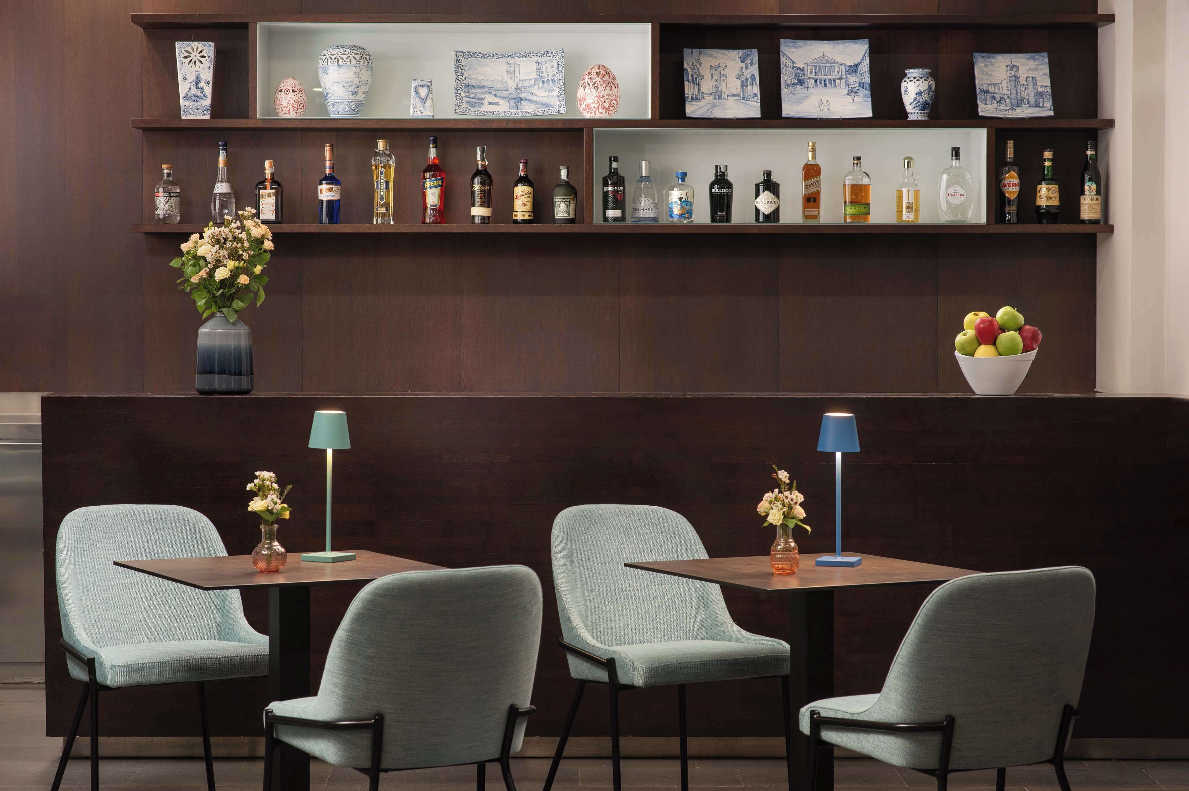 A bar area with multiple liquor bottles, a vase with flowers, and a bowl of fruit. There are two tables with chairs and two floor lamps.