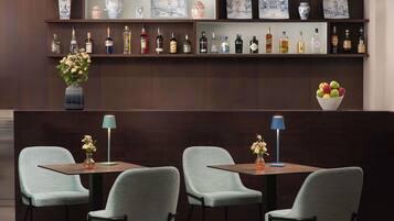 A bar area with multiple liquor bottles, a vase with flowers, and a bowl of fruit. There are two tables with chairs and two floor lamps.