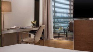 A modern hotel room with a bed, a desk with a lamp, a chair, a TV, and a view of the city through large windows.