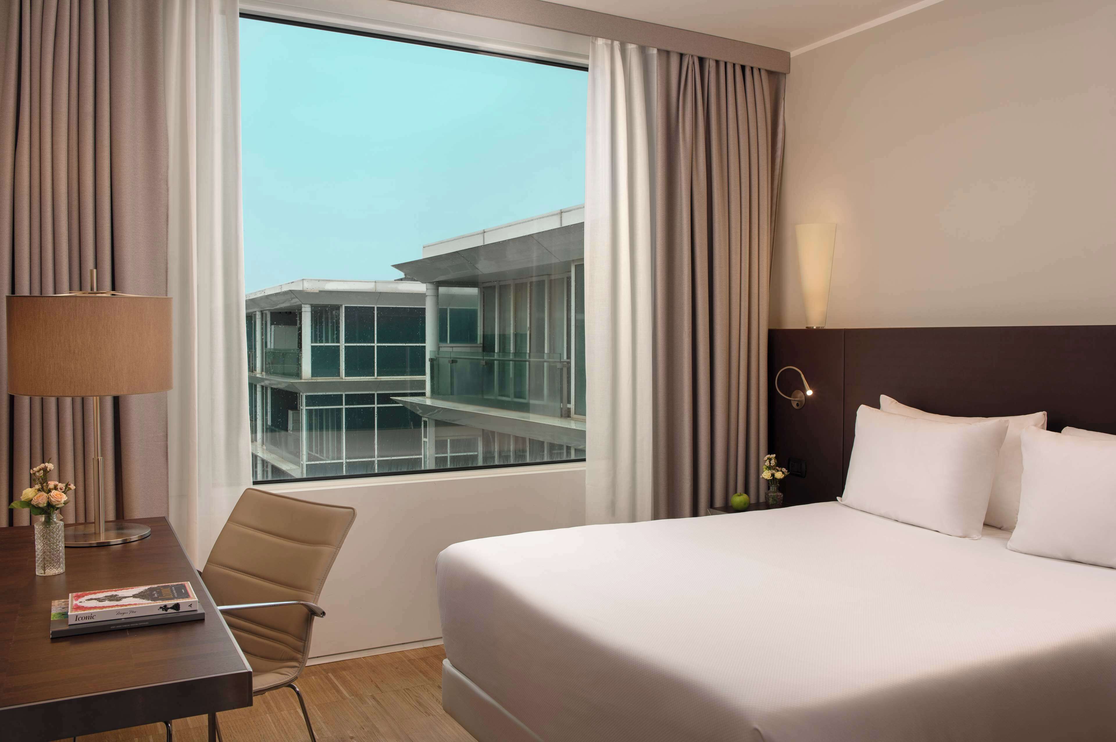 A hotel room with a large bed, a desk with a lamp, and a view of a modern building through the window.