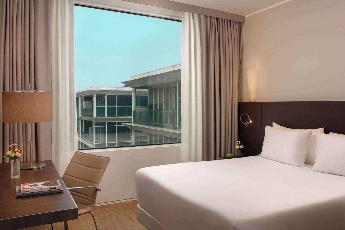 A hotel room with a large bed, a desk with a lamp, and a view of a modern building through the window.
