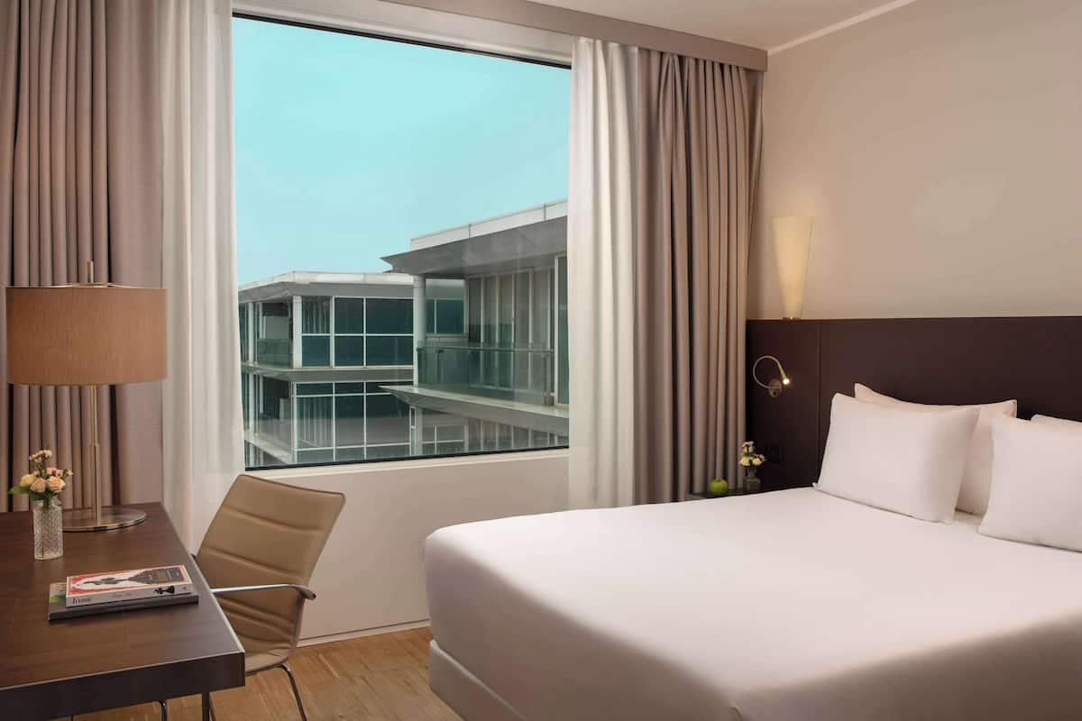 A hotel room with a large bed, a desk with a lamp, and a view of a modern building through the window.