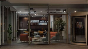 A modern hotel lobby with glass walls, comfortable seating, a bar area, and a visible plant.