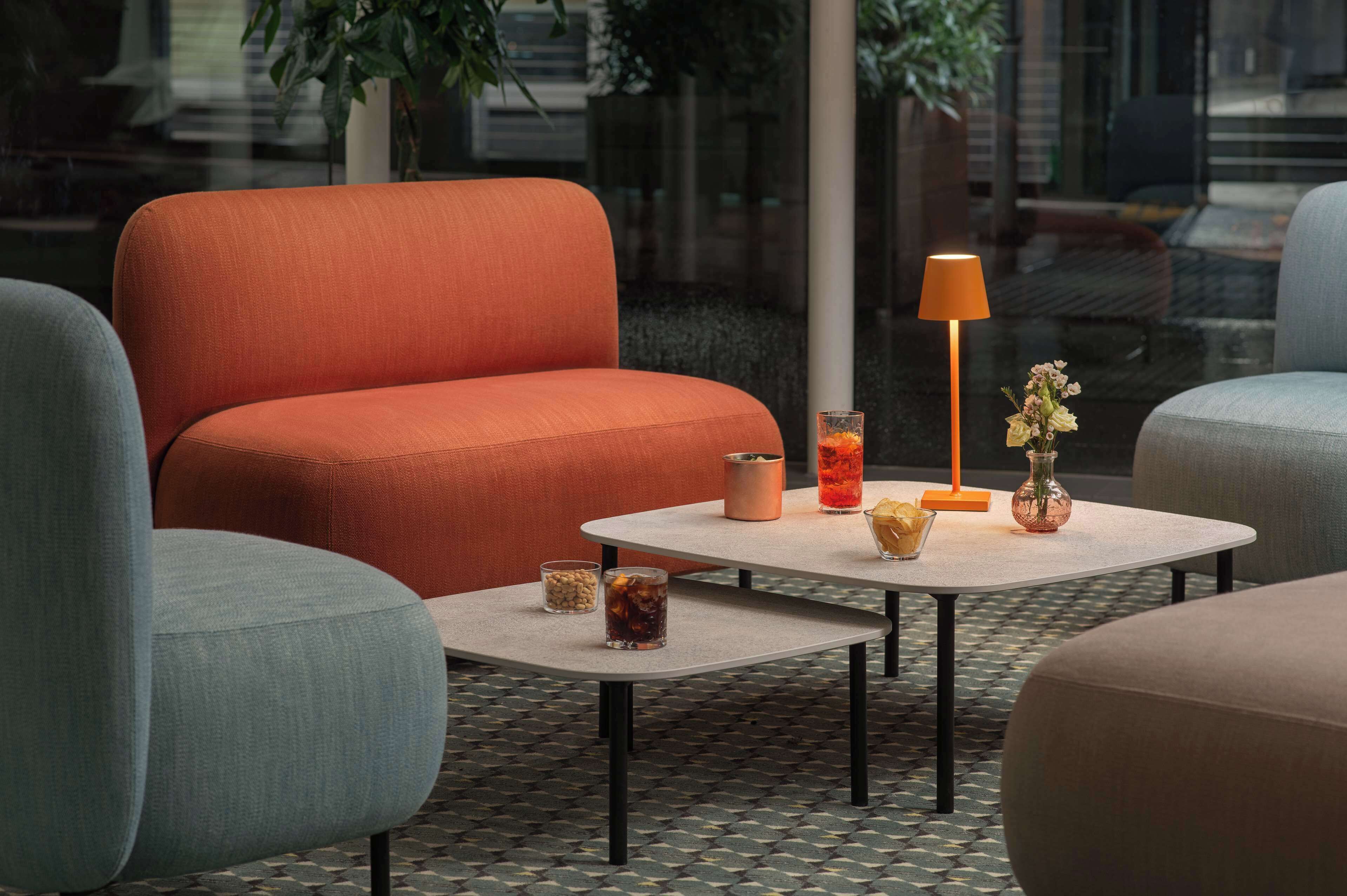 A modern lounge area with a sofa, armchairs, a small table with drinks, a lamp, and a vase with flowers.