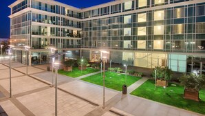 A modern building with multiple floors, large glass windows, and a well-lit outdoor area with planters and a paved walkway.