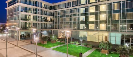 A modern building with multiple floors, large glass windows, and a well-lit outdoor area with planters and a paved walkway.