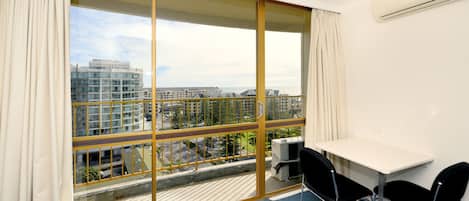 Quarto de hotel moderno com uma grande porta de vidro deslizante, uma escrivaninha branca com duas cadeiras pretas e vista para edifícios urbanos.