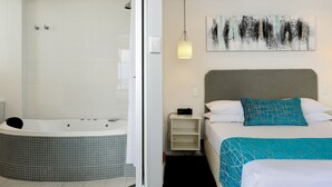 A modern bathroom with a bathtub and shower, a bedside table with a lamp, and a bed with a headboard and decorative pillows.