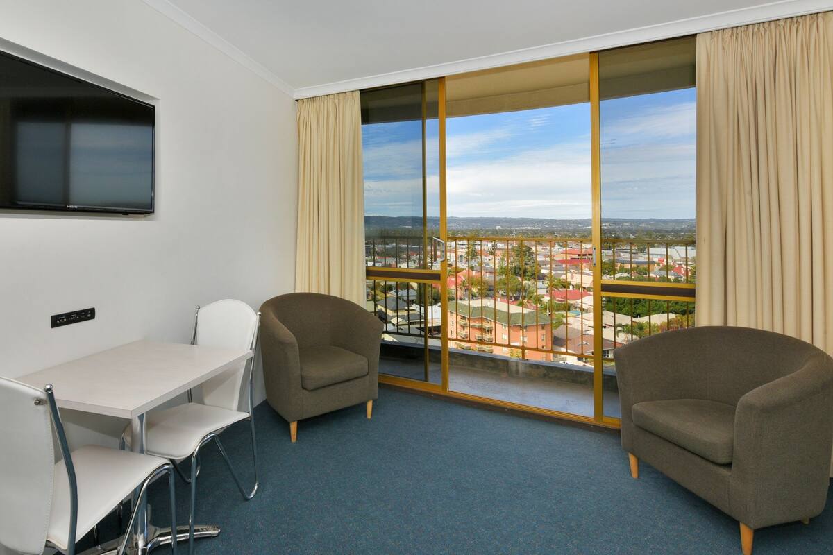 Ett hotellrum med en balkong, en tv som är monterad på väggen, ett skrivbord med stolar och en soffa.