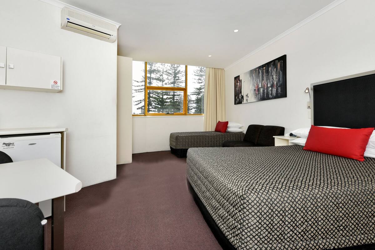 Ett hotellrum med en säng, ett skrivbord, en stol, ett fönster med utsikt över träd och en luftkonditioneringsanläggning.