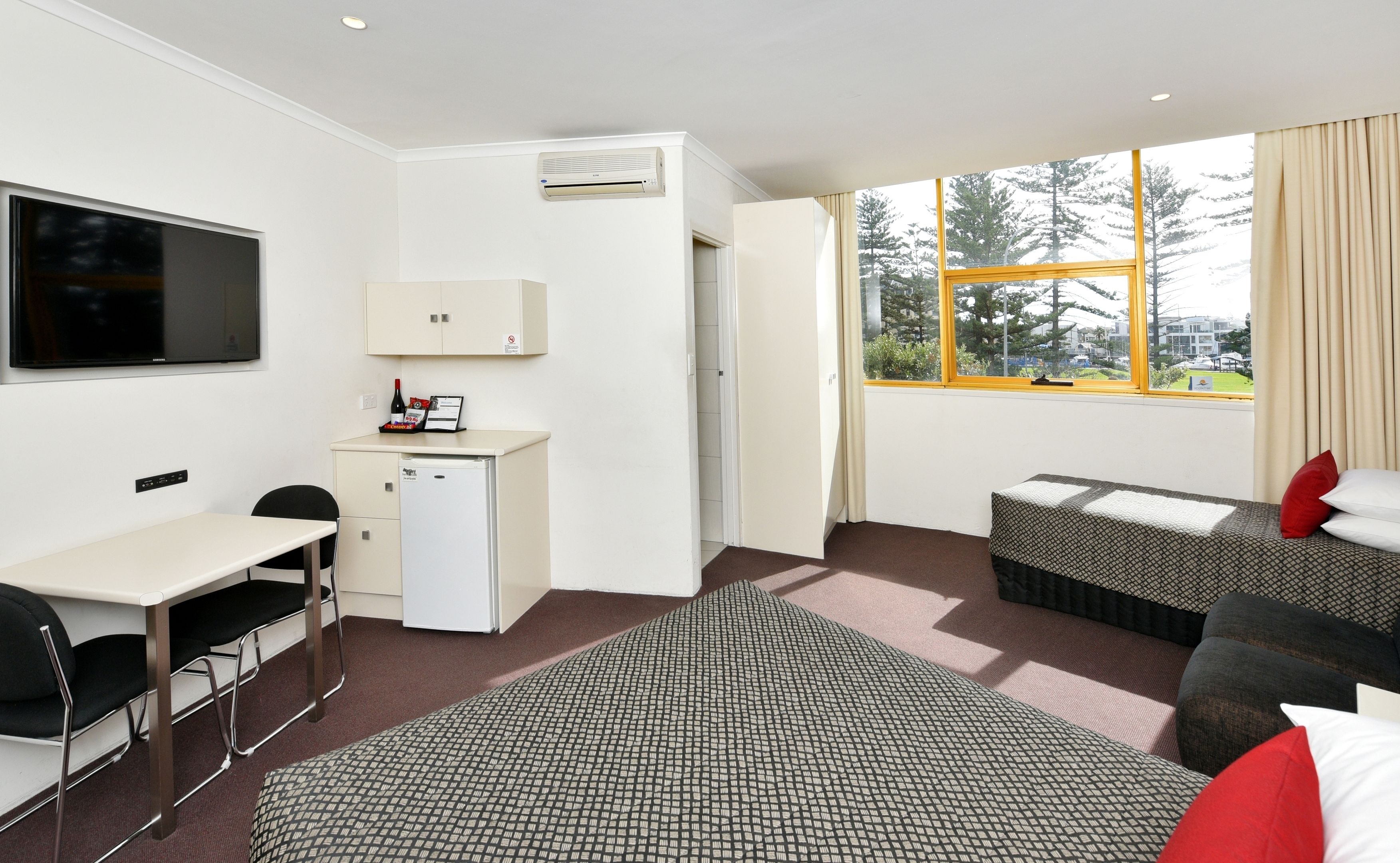 Habitación de hotel con escritorio, un refrigerador pequeño, televisor, dos camas y ventana con vista a árboles.