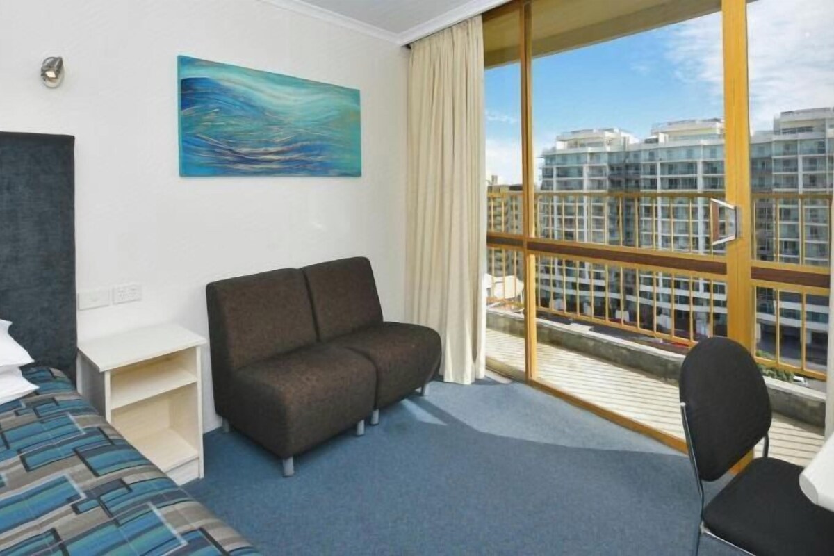 Habitación de hotel con cama, sofá, escritorio y silla. Amplia ventana con vistas a edificios.