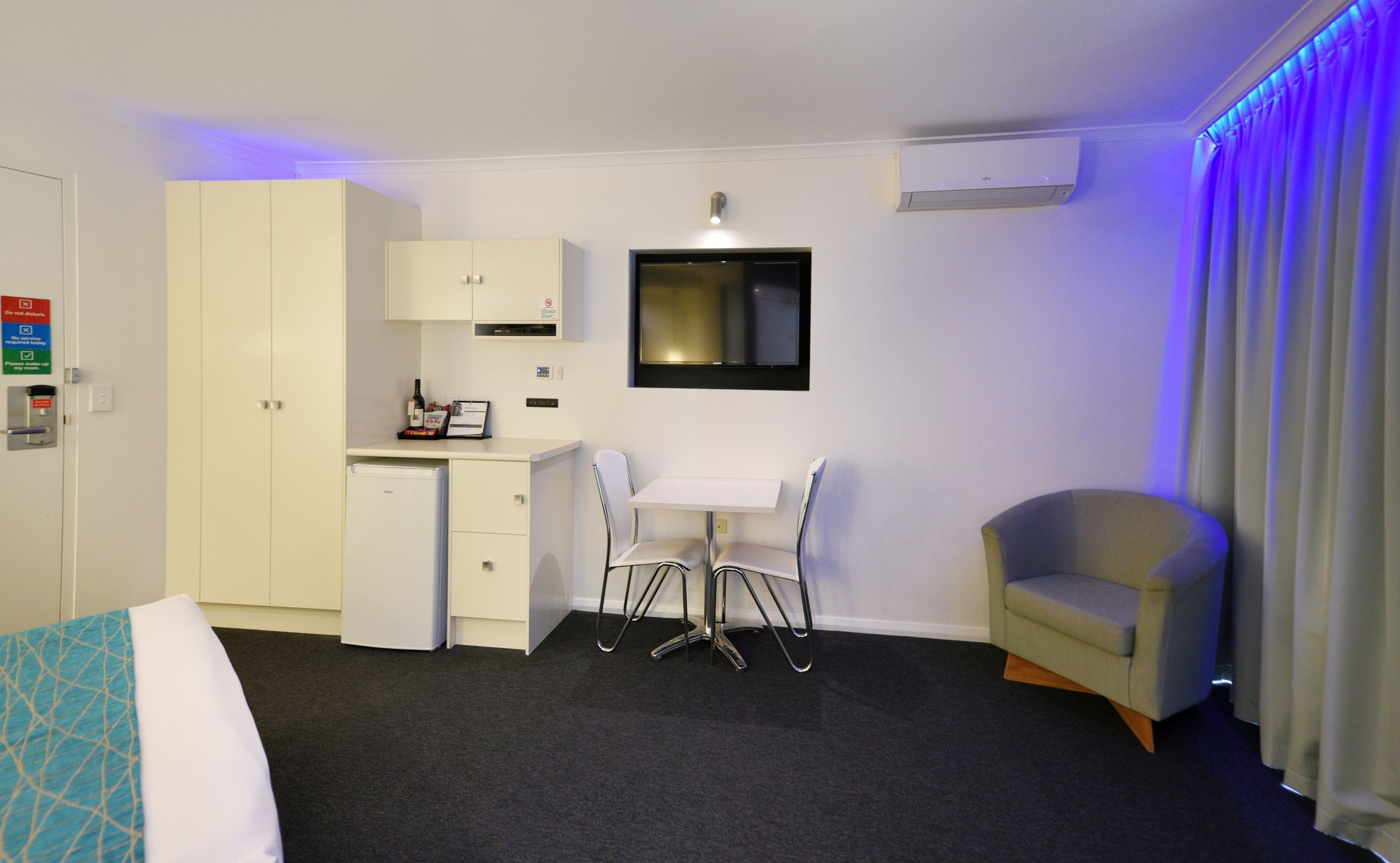 Une chambre d’hôtel comprenant un lit, un bureau avec deux chaises, un petit coin cuisine, une télévision et une fenêtre avec des rideaux.