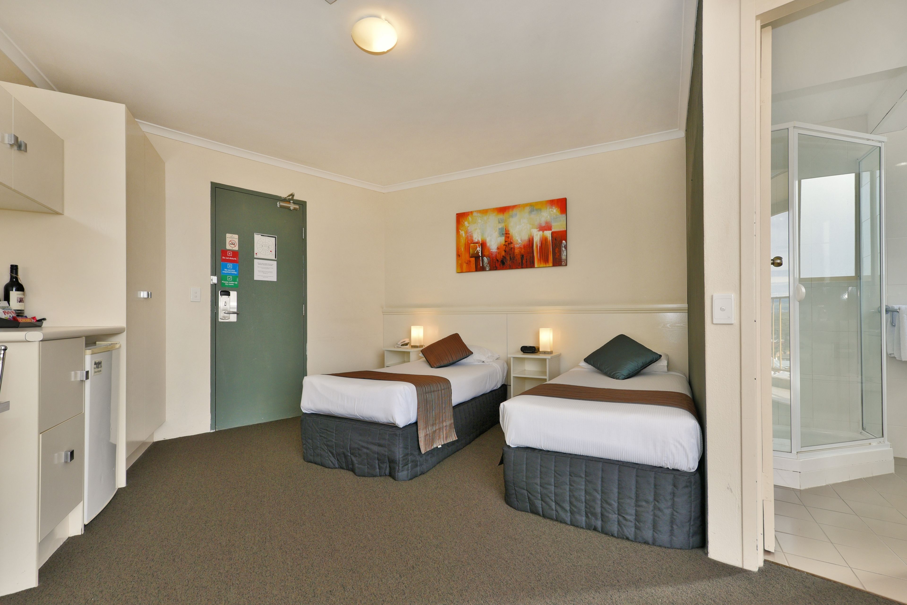 A hotel room with two beds, a desk, a small fridge, and a shower area.