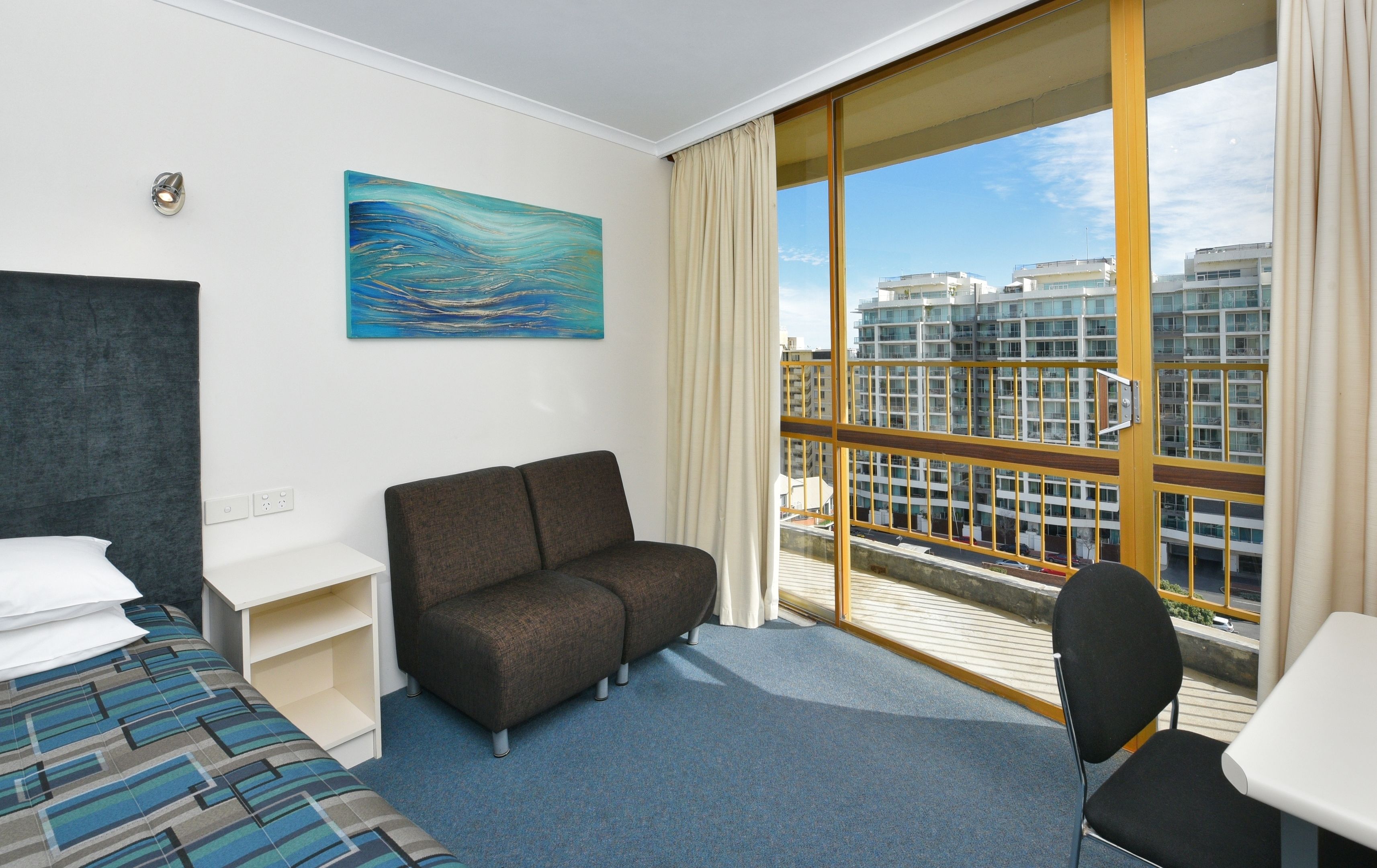 A hotel room with a bed, a sofa, a desk, and a chair. There is a large window with a view of buildings.