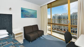 A hotel room with a bed, a sofa, a desk, and a chair. There is a large window with a view of buildings.