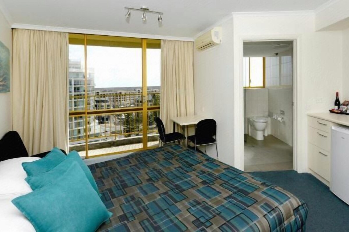 A hotel room with a large bed, a desk, a chair, and a view of the city through large windows.
