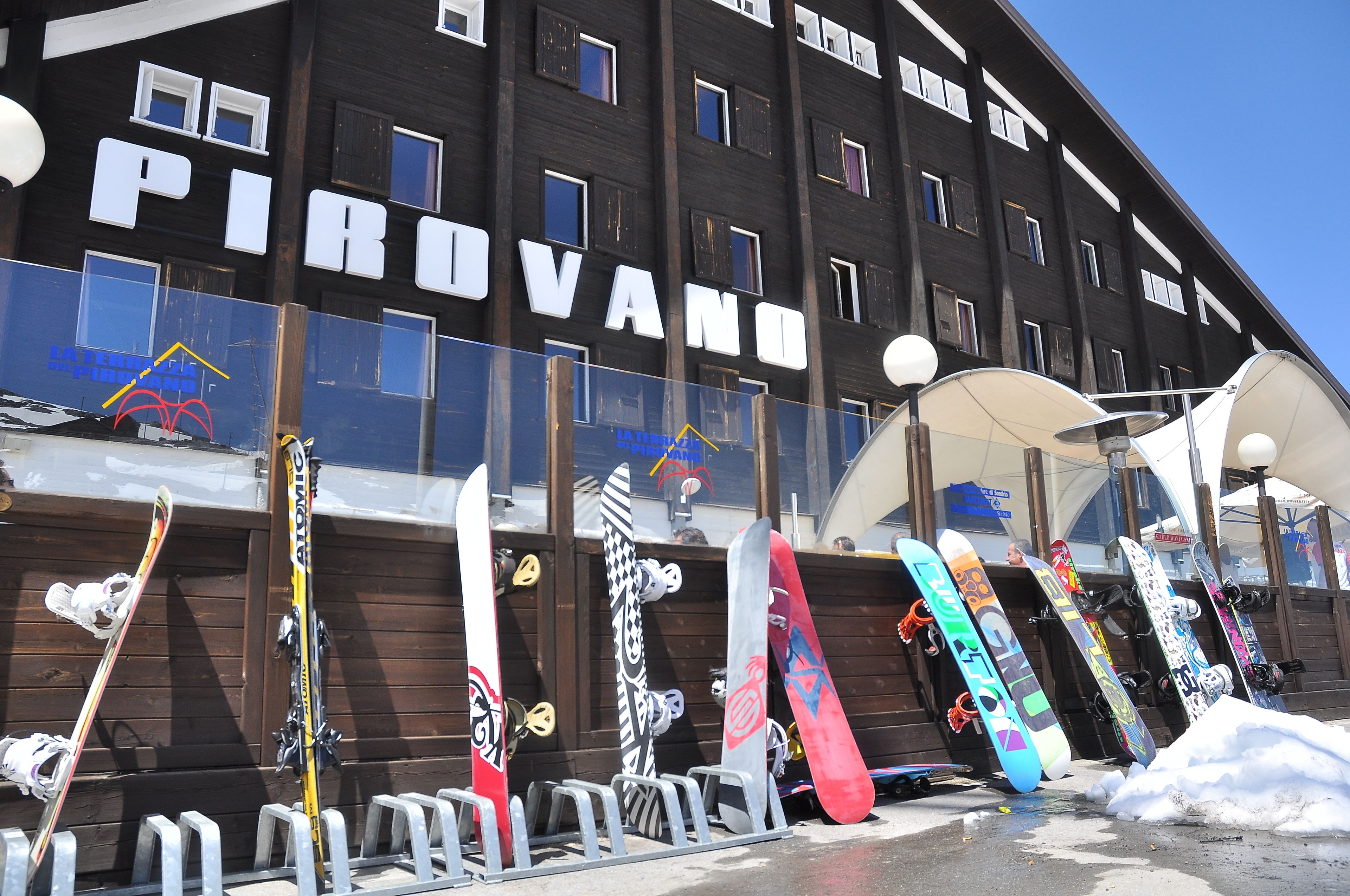 A building with the name "PIROVANO" and a row of snowboards leaning against a railing outside.