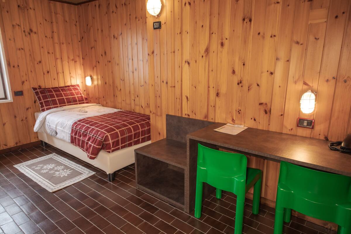 Ein Zimmer mit Holzwänden, einem Bett, einem Schreibtisch und zwei grünen Stühlen.