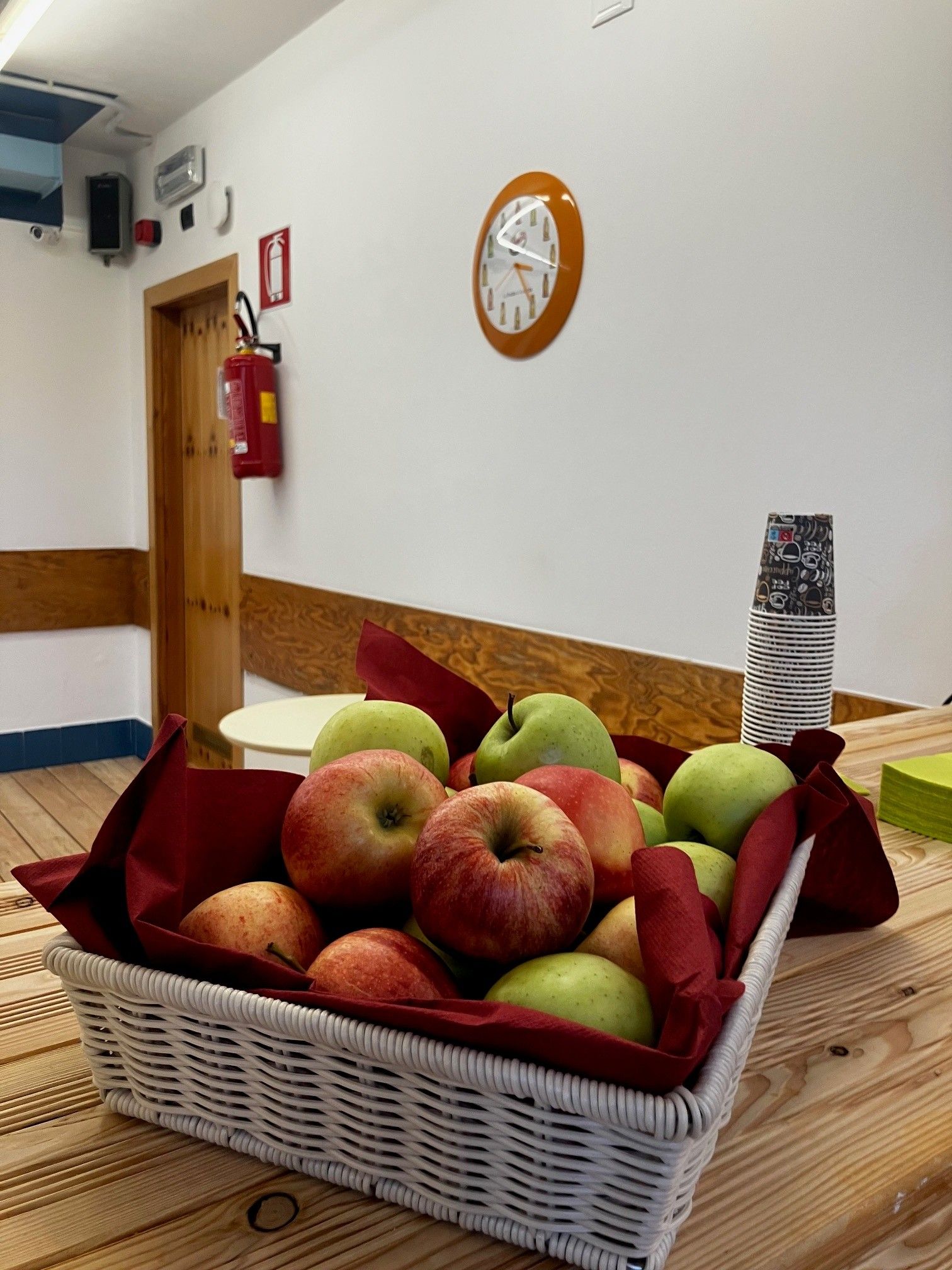 Una cesta de mimbre llena de manzanas sobre un suelo de madera, un extintor, un reloj y una pared con diseño.