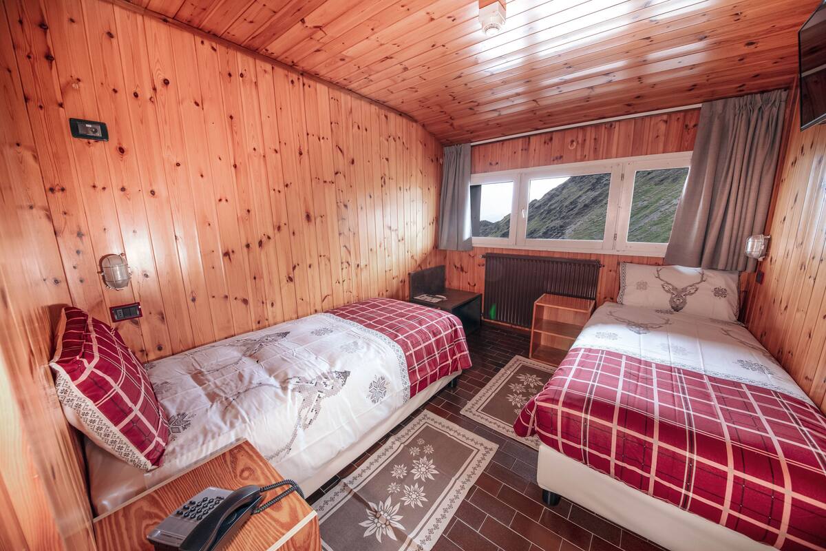 Eine Holzhütte mit zwei Betten, einem kleinen Tisch, einem Stuhl, einem Heizgerät und einem Fenster mit Blick auf Hügel.