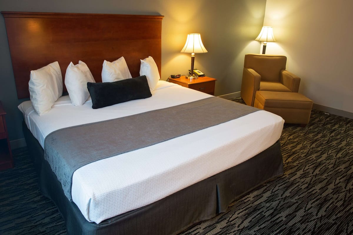 A hotel room with a large bed, a wooden headboard, two bedside lamps, a chair, and a carpeted floor.