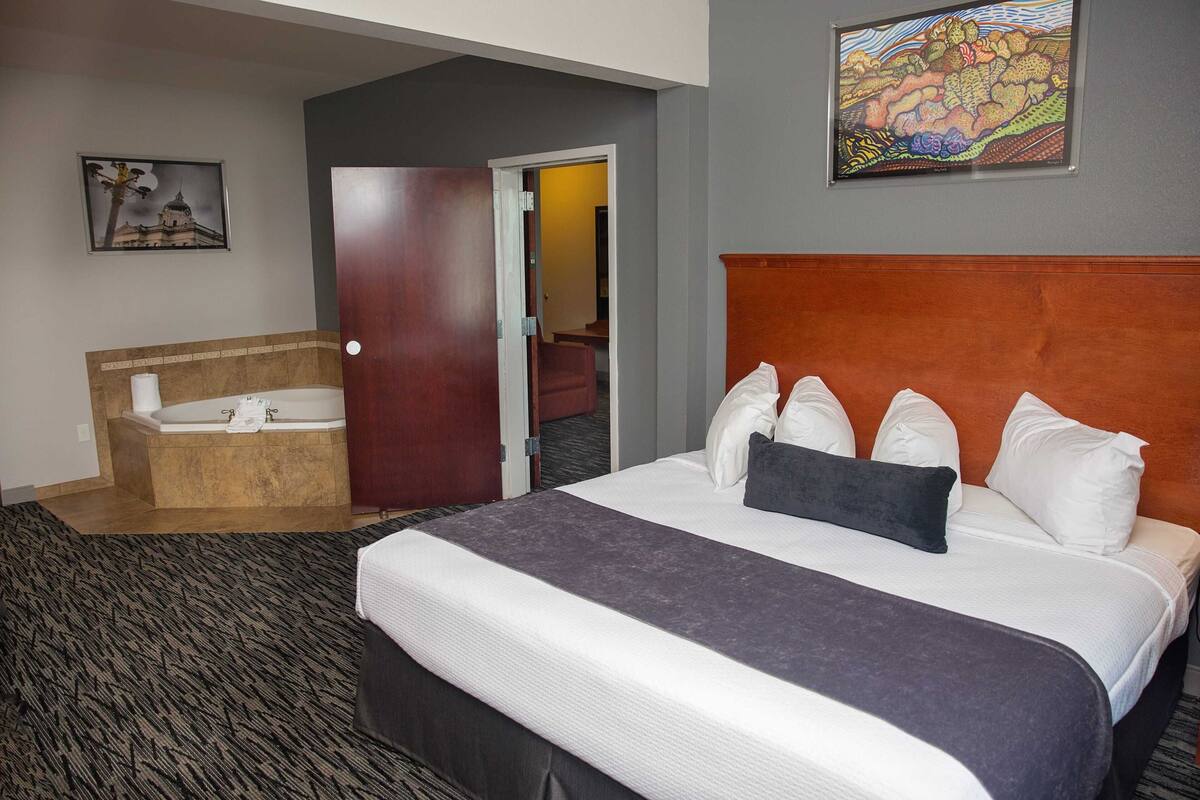 A hotel room with a large bed, a separate bathtub, a door leading to another room, and a painting on the wall.