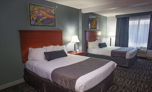 Quarto de hotel com duas camas, uma mesinha de cabeceira, uma luminária, um quadro na parede e uma janela com cortinas.