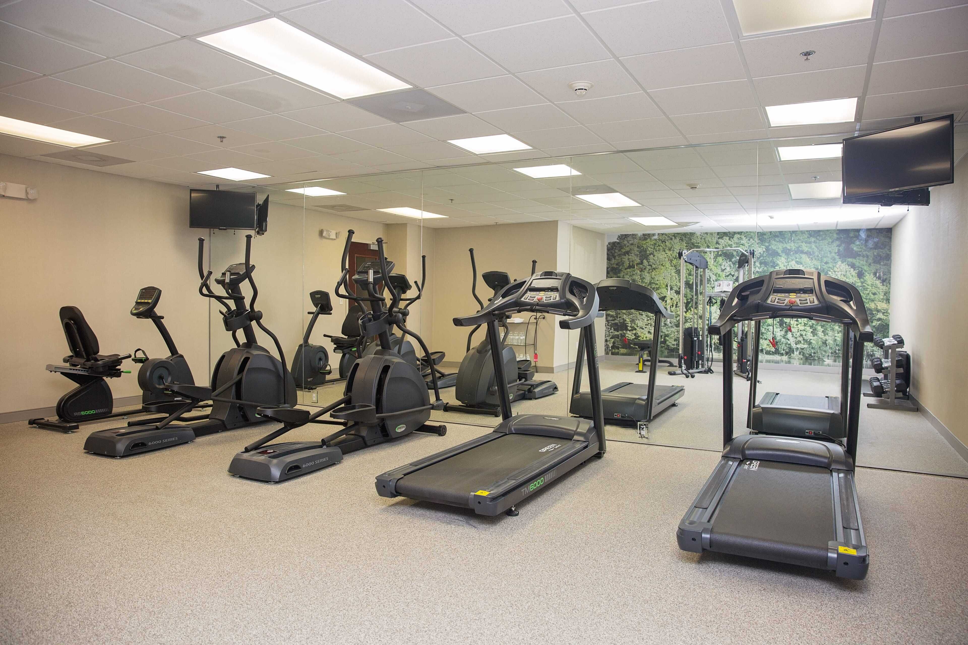 Un gimnasio con diversas máquinas de cardio, un espejo grande en la pared y un televisor montado.
