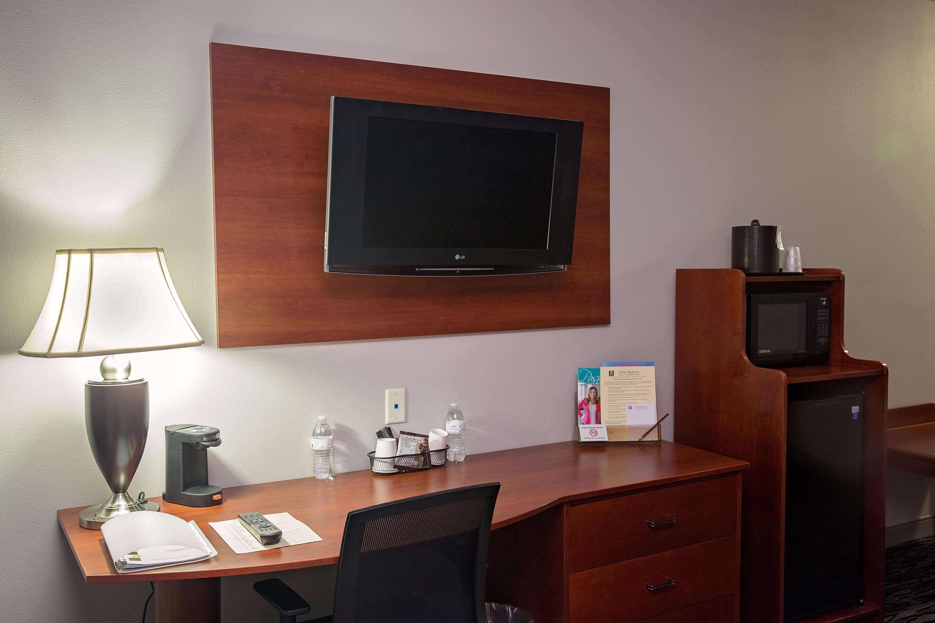 Habitación de hotel con televisión de pantalla plana en la pared, un escritorio con lámpara, cafetera, botellas de agua, control remoto y refrigerador.