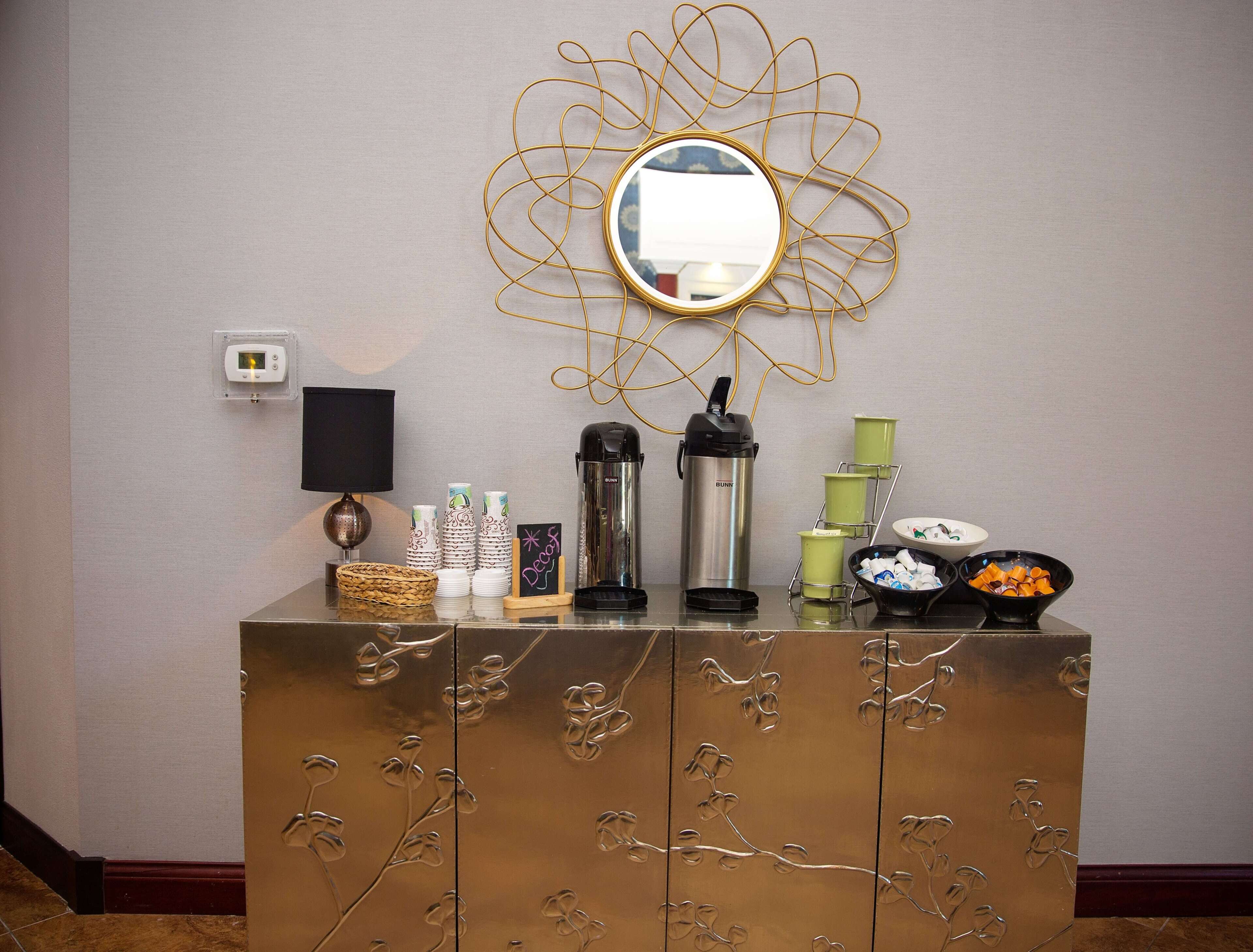 A room with a decorative mirror, a coffee maker, a thermos, and a bowl of fruit on a counter.