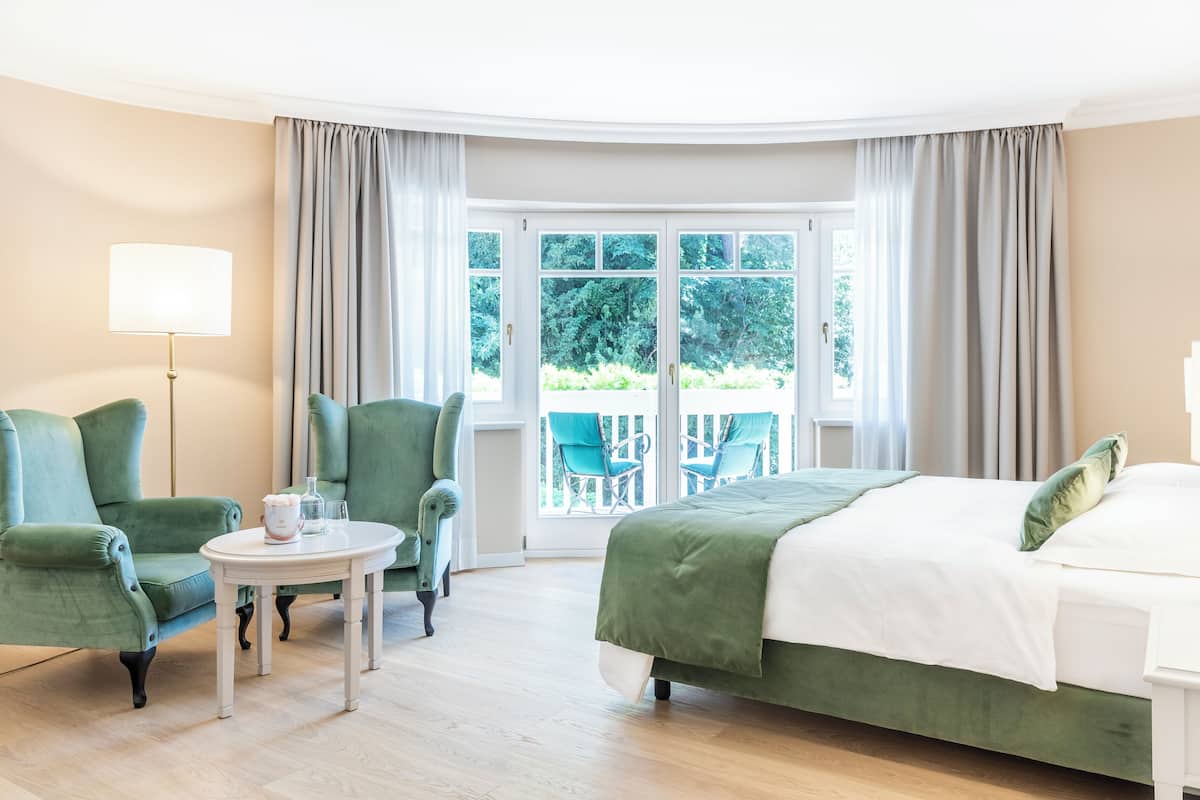 Habitación de hotel con una cama, dos sillones verdes, una mesita de noche con lámpara y vistas a zonas verdes a través de amplias ventanas.