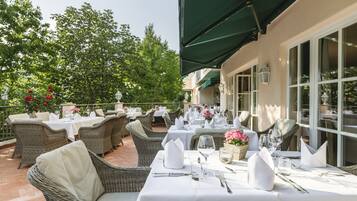 Un área de comedor al aire libre con mobiliario de mimbre y manteles blancos, rodeada de follaje y ventanas grandes.