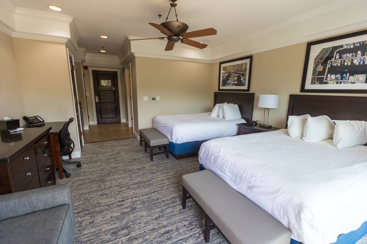 Ein Hotelzimmer mit zwei Betten, einem Schreibtisch, einem Stuhl, einer Bank, einem Deckenventilator und einem gerahmten Bild an der Wand.