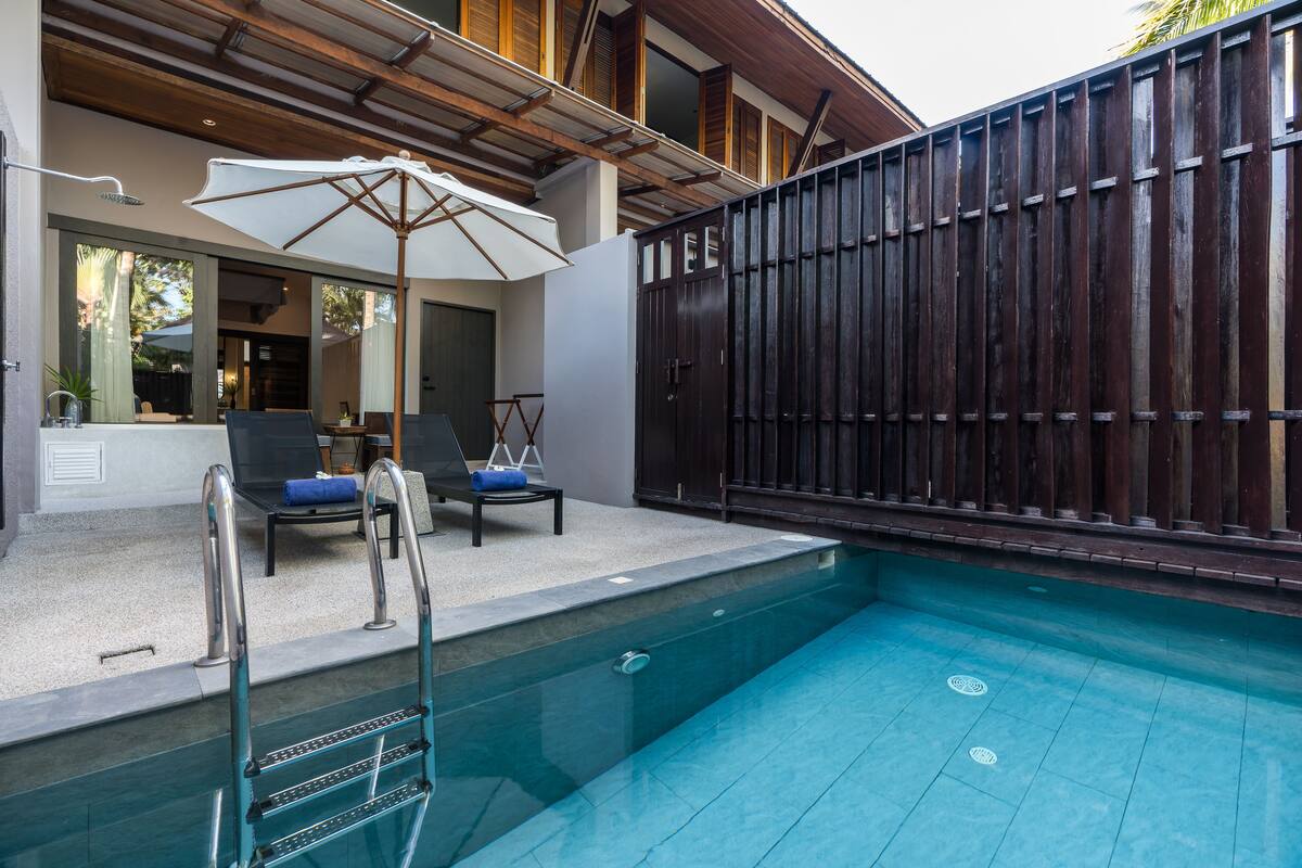 Una piscina con valla de madera, sillas de descanso y un área de estar cubierta.