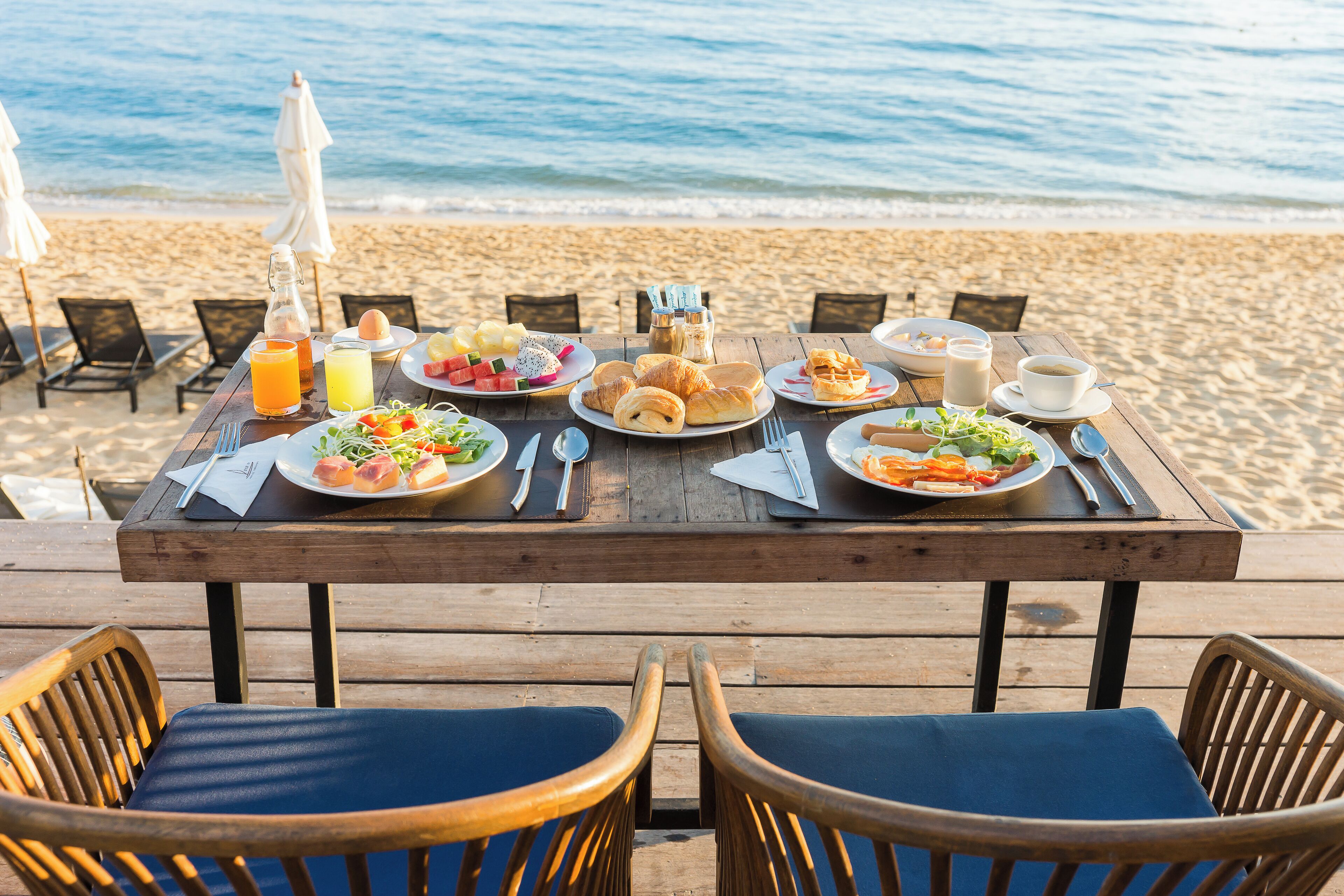 Una mesa de madera dispuesta con alimentos para el desayuno frente a una playa con vista al océano.