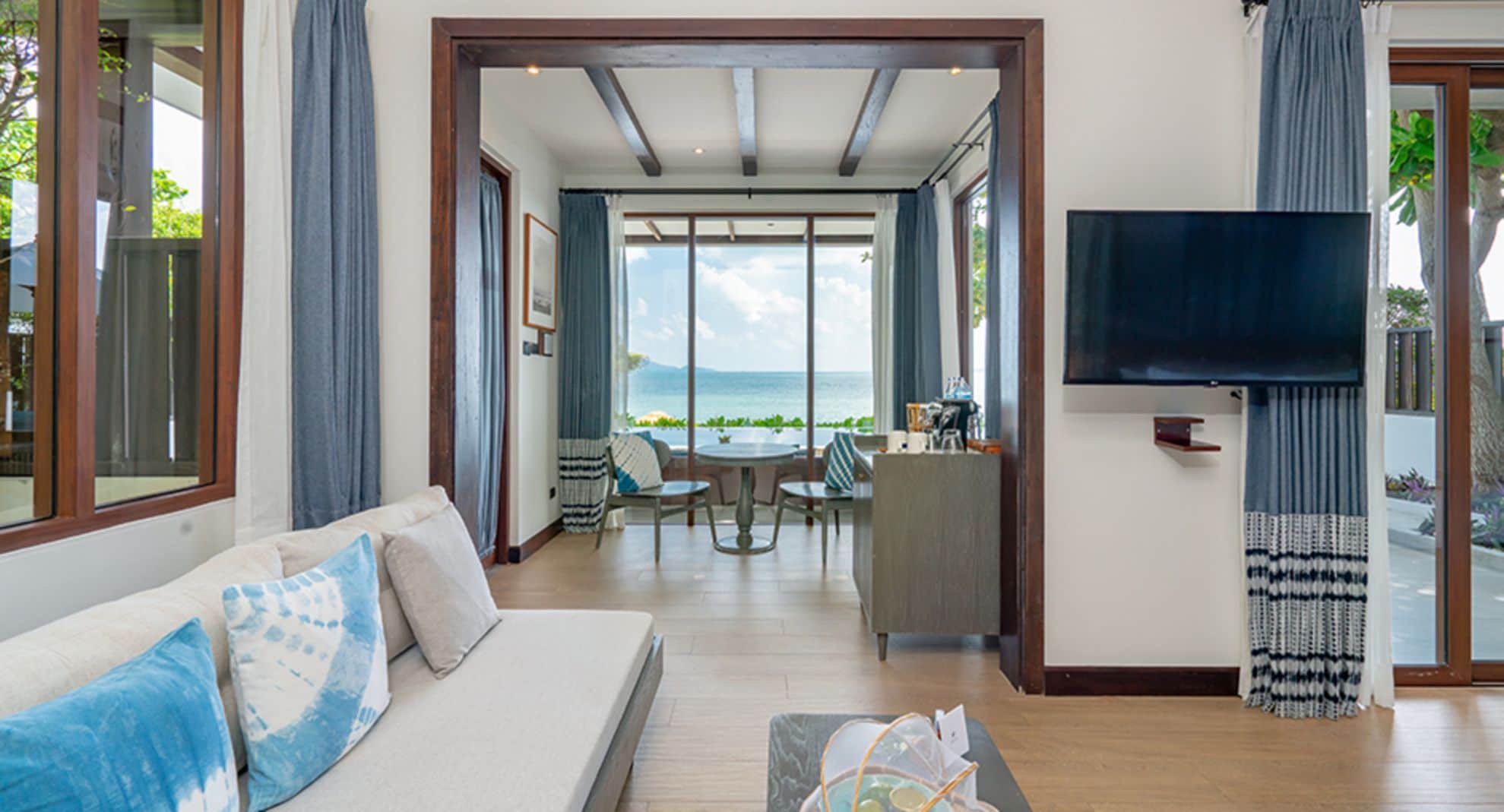 Uma sala de estar moderna com sofá, área de jantar com mesa e cadeiras, TV montada na parede e vista para o mar através de grandes janelas.