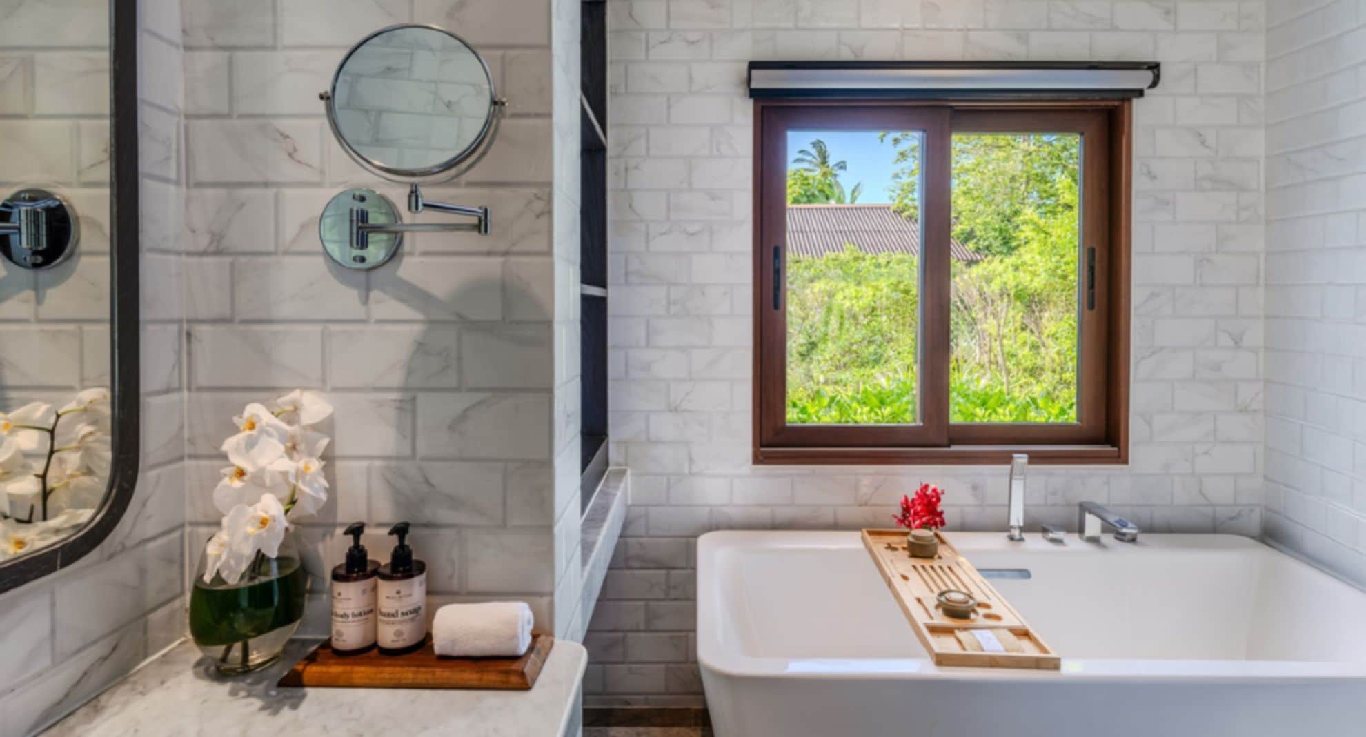 Un baño con bañera, ventana con vistas, espejo y estante con artículos de aseo.