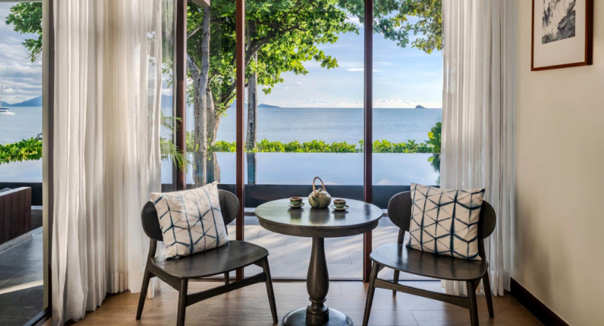 Una habitación con vistas al mar, dos sillas con cojines estampados, una mesita con tetera y tazas, y cortinas translúcidas.