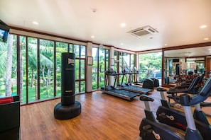 A gym with wooden flooring, exercise equipment, and large windows offering a view of greenery. - NH Collection Samui Peace Resort (Koh Samui)