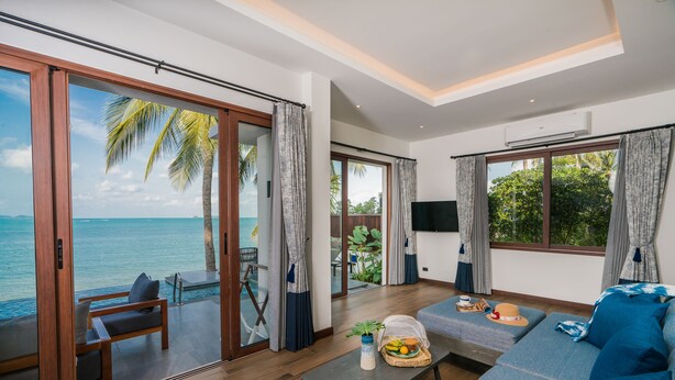 A modern living room with a large sliding glass door leading to a balcony with a view of the ocean. There is a sofa, a coffee table, and a TV mounted on the wall. - NH Collection Samui Peace Resort (Koh Samui)
