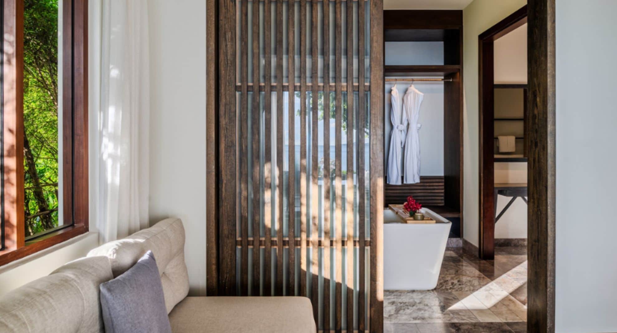 A modern hotel room with a sofa, a wooden partition, a view of a pool, and a bathroom with a bathtub and towels.