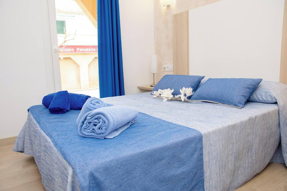 Un lit bien fait, avec du linge de lit bleu et des oreillers, une petite table de chevet avec une lampe, et une vue sur un bâtiment à l’extérieur, visible à travers une fenêtre ornée de rideaux bleus.