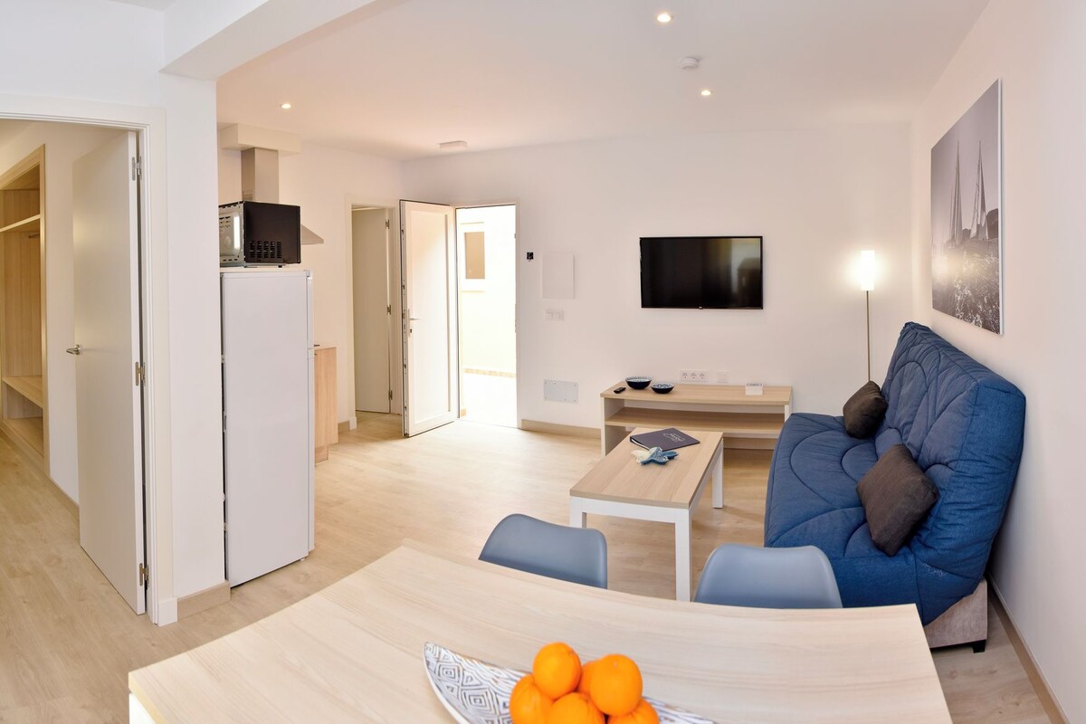 Un salon moderne avec un canapé bleu, une table à manger sur laquelle se trouve un saladier rempli de fruits, et un téléviseur fixé au mur.